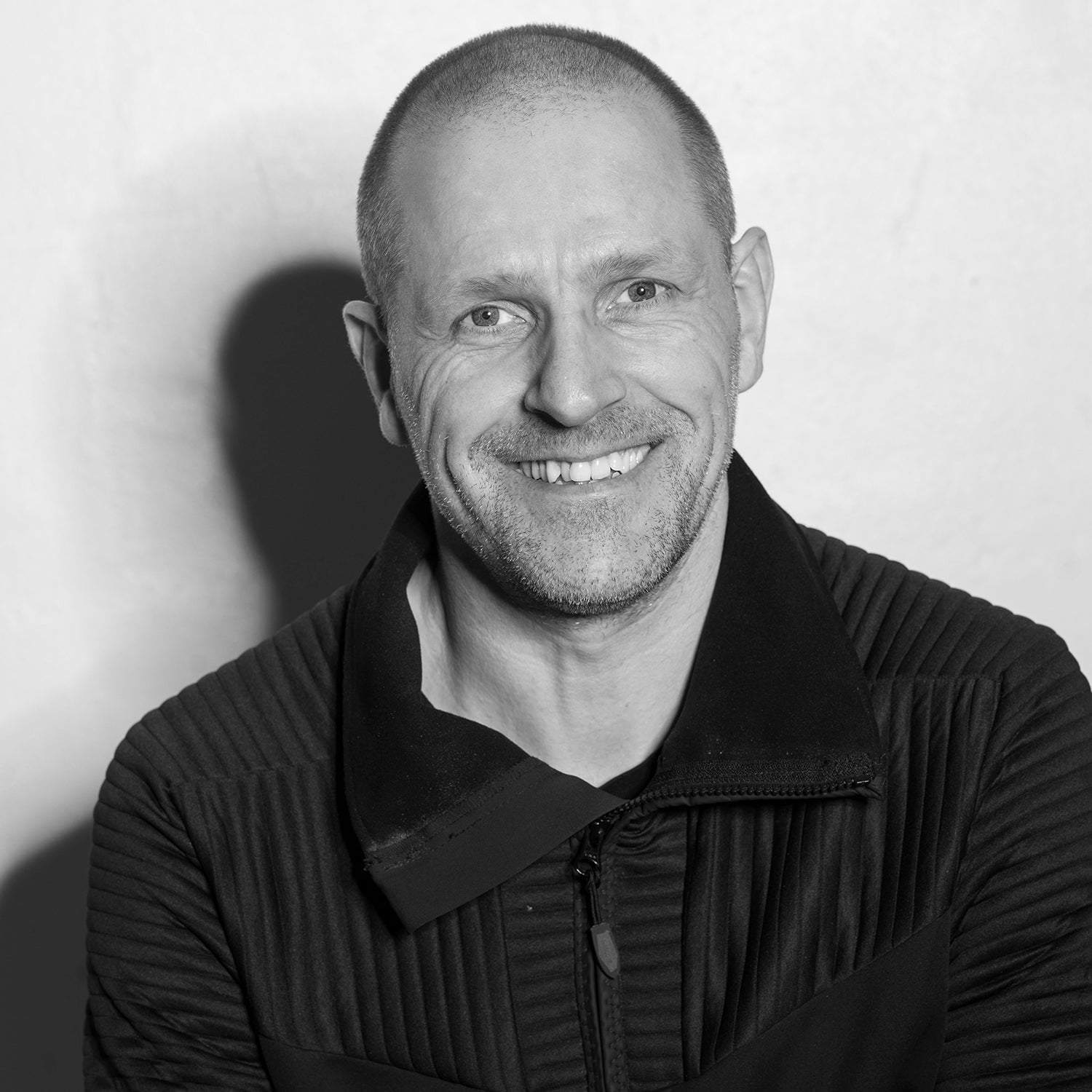 Mann mit kahl geschorenem Kopf lächelt in die Kamera, trägt schwarze Jacke.
