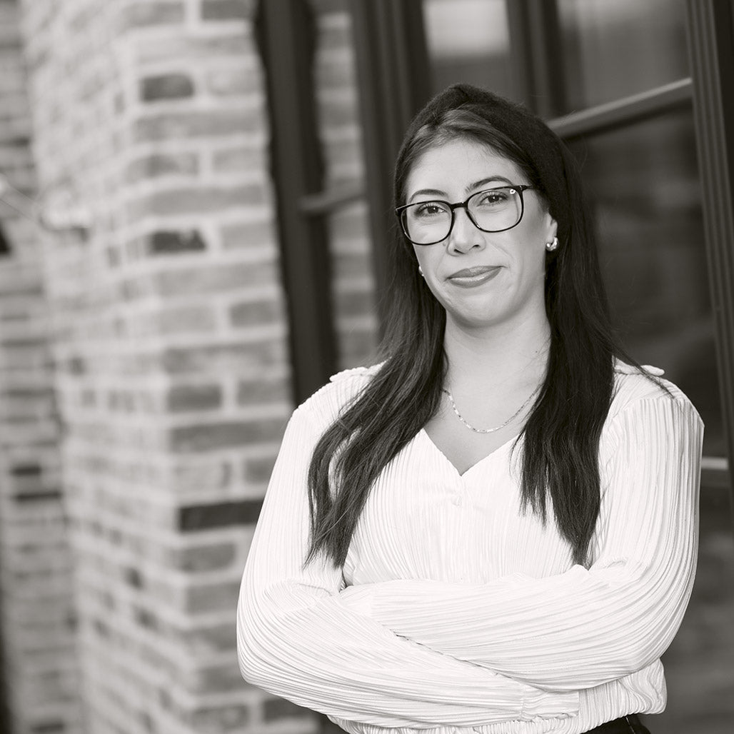 Frau mit langer Brille, Haare offen, weißes Oberteil, vor Backsteinmauer stehend.