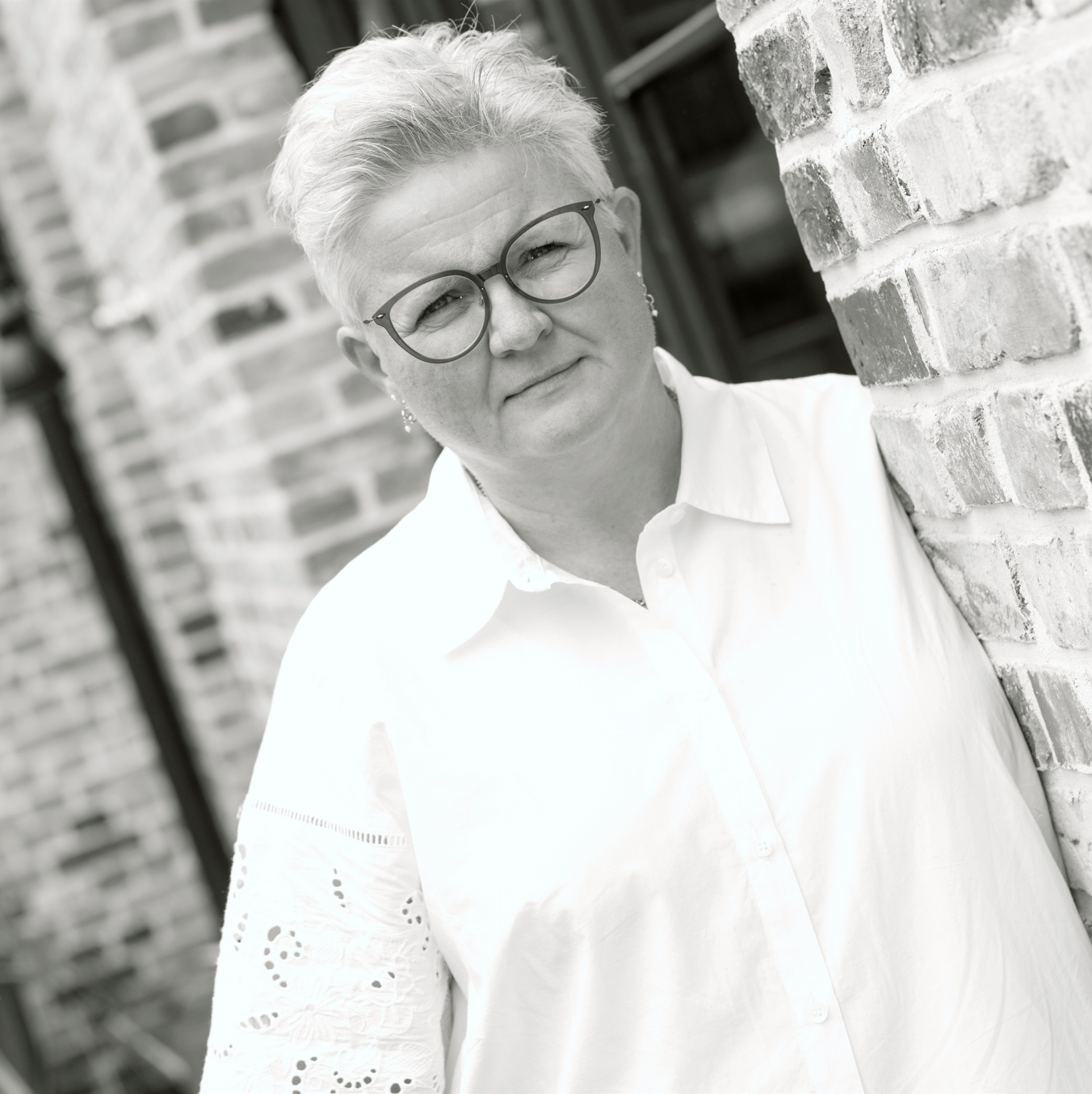 Frau mit kurzen Haaren und Brille lehnt an einer Ziegelwand.