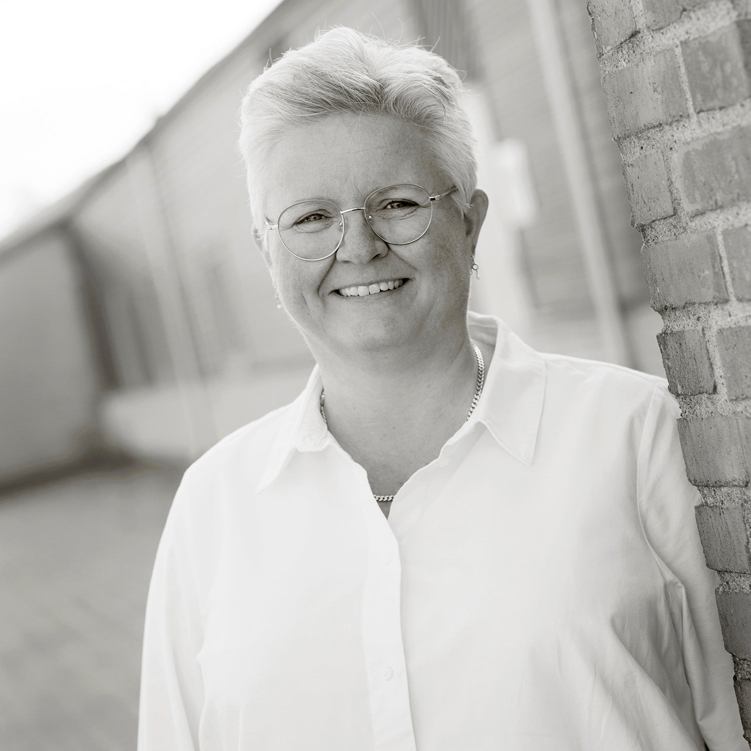 Frau mit kurzem Haar und Brille in weißem Hemd lächelt in die Kamera, an Ziegelwand stehend.