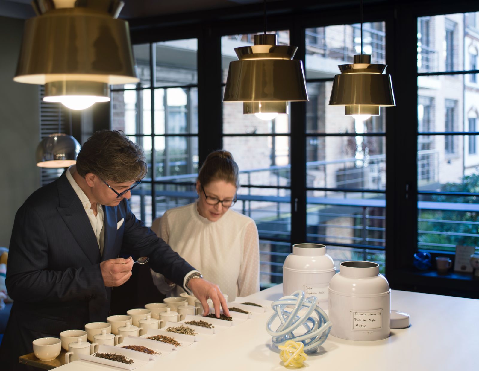 Zwei Personen stehen an einem Tisch mit Teeproben unter goldenen Lampen.