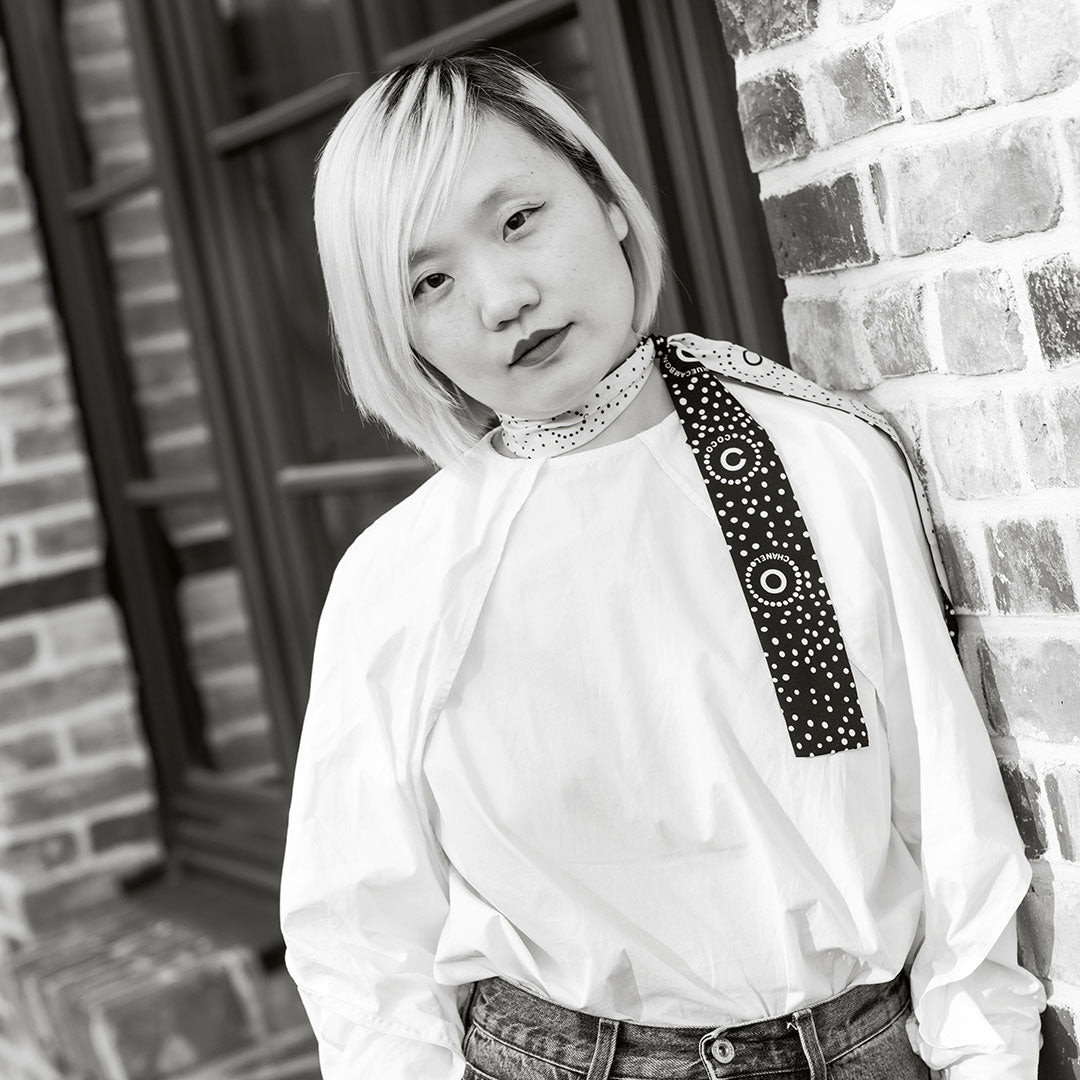 Person mit kurzem, hellem Haar trägt ein weißes Oberteil und steht vor einer Mauer.