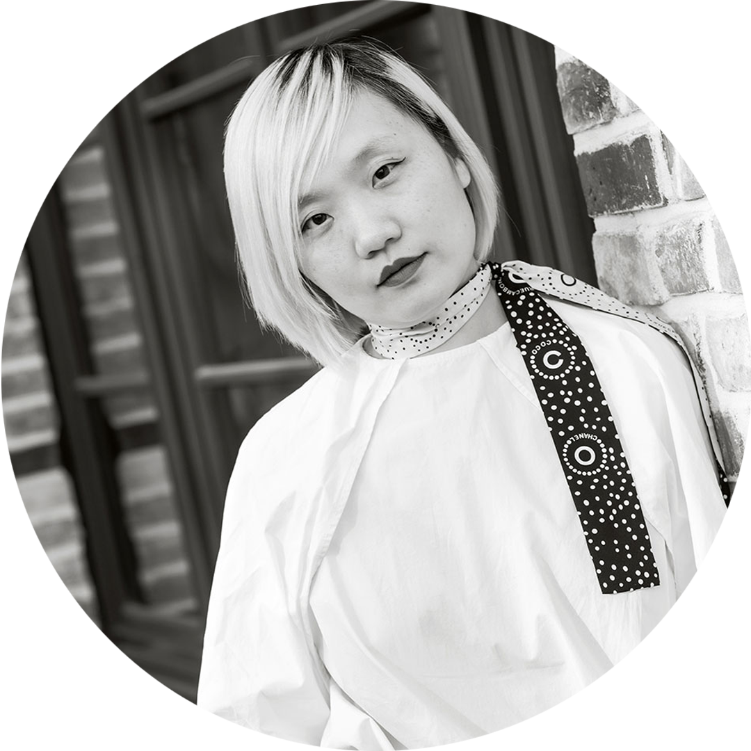 Schwarz-weiß-Porträt einer Person mit hellem Haar und weißem Oberteil, lehnt an Ziegelwand.